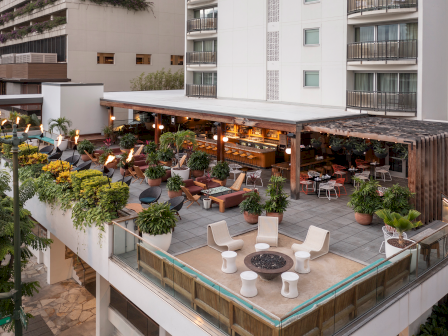 The image shows a rooftop terrace with seating areas, plants, and a bar near a building, offering an inviting outdoor space.