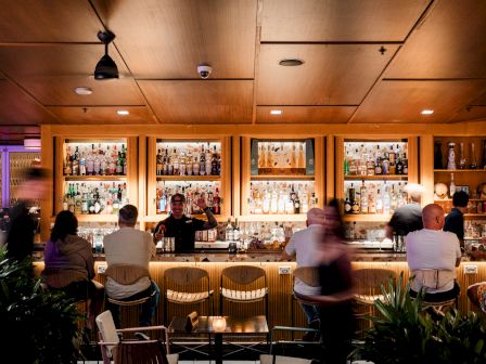 A cozy bar scene with warm lighting, colleagues or friends seated at a wooden counter enjoying drinks, shelves stocked with bottles behind.
