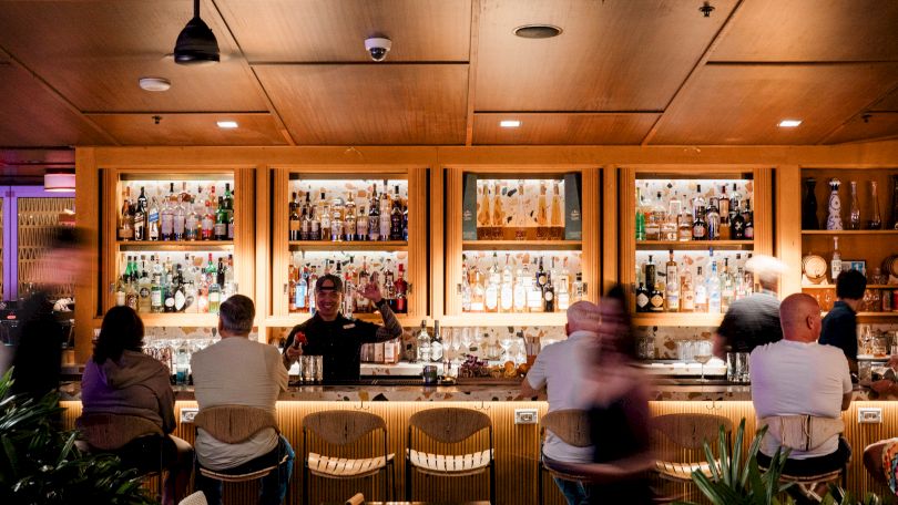 A cozy bar scene with warm lighting, colleagues or friends seated at a wooden counter enjoying drinks, shelves stocked with bottles behind.
