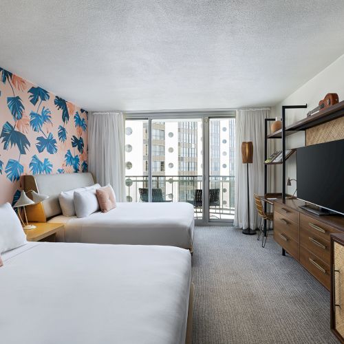 A modern hotel room with two beds, a colorful palm‑print accent wall, a desk and TV stand, and a balcony view of the city, all in warm, neutral tones.