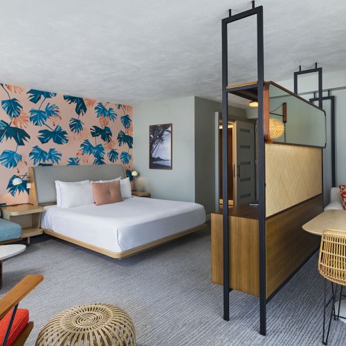 A bright hotel suite with a blue palm-printed accent wall, a white bed, a teal sofa, mid-century chairs, and a partitioned seating area by a small dining table.