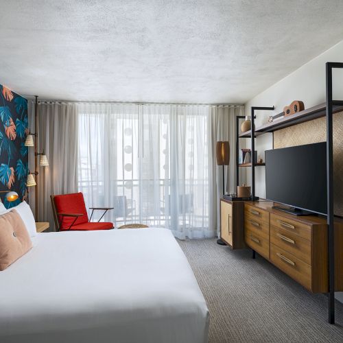 A modern hotel room with a tropical flower wallpaper accent wall, a large bed with white linens, a red chair, a wooden dresser with a TV, and a full-length mirror.