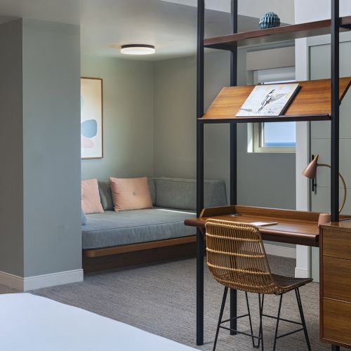A modern hotel room with a study desk, a woven chair, and a built-in bench seating area with cushions, plus wall art and soft lighting.