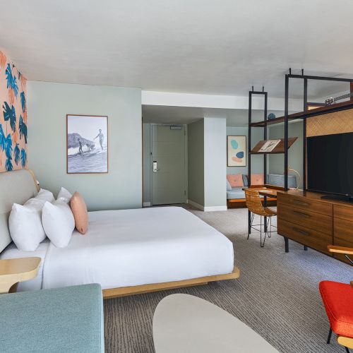 A bright hotel room with a white bed, tropical blue leaf wallpaper, a teal sofa, wooden desk and TV unit, red chair, and open shelving in a modern, airy layout.