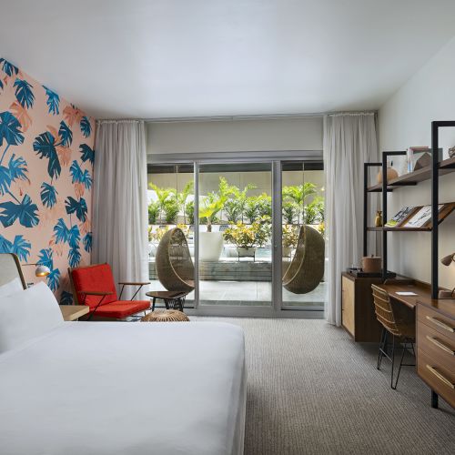 A modern hotel room with a bold blue palm wallpaper feature wall, two white beds, a red chair, hanging chairs by the balcony, and a wooden desk and dresser facing a flat-screen TV, ending here.