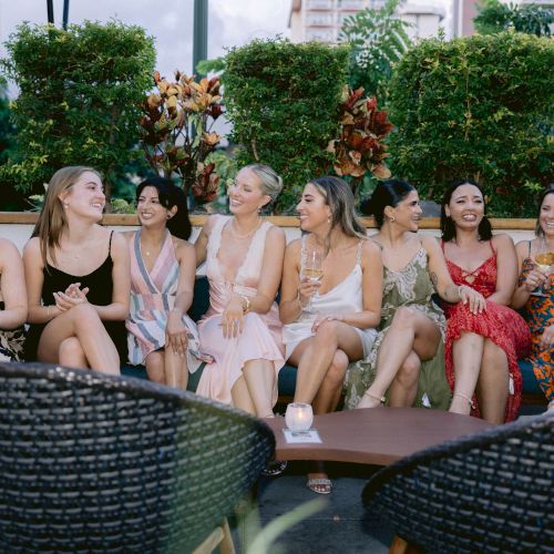 A group of people posing outdoors, some in swimwear, with a dog on a couch and greenery in the background.