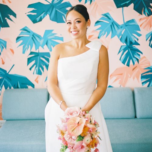 A smiling bride in a white one-shoulder gown holds a pink-peach bouquet, posing against a tropical leaf mural with a blue sofa backdrop.