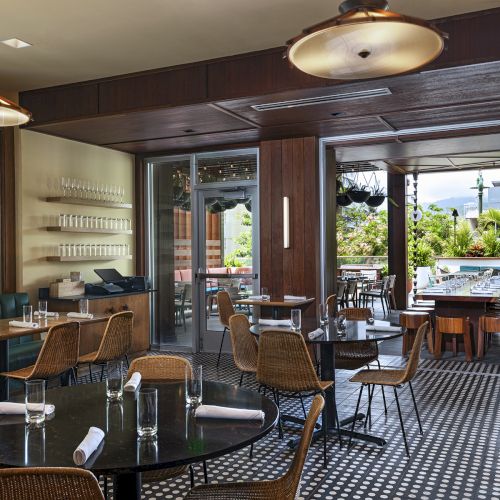 A stylish, airy café with wicker chairs, round tables, a teal banquette, oak counters, and large windows opening to a sunny outdoor patio.