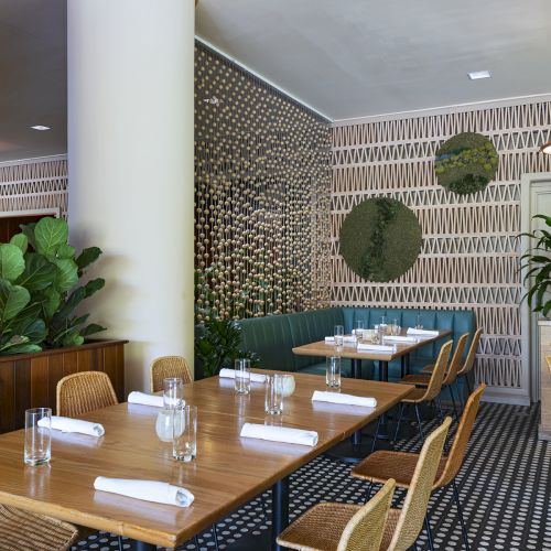 A stylish restaurant interior with wooden tables, woven chairs, potted plants, a patterned wall, and a teal booth along a pillar.