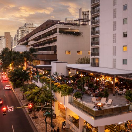 A bustling city street at dusk with tall buildings, storefronts, and cars; warm lights glow from shops while pedestrians stroll along the sidewalk.