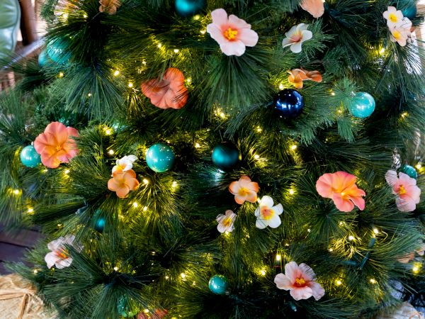 A decorated Christmas tree with green tinsel, warm lights, teal and blue ornaments, and pink/orange flower decorations.