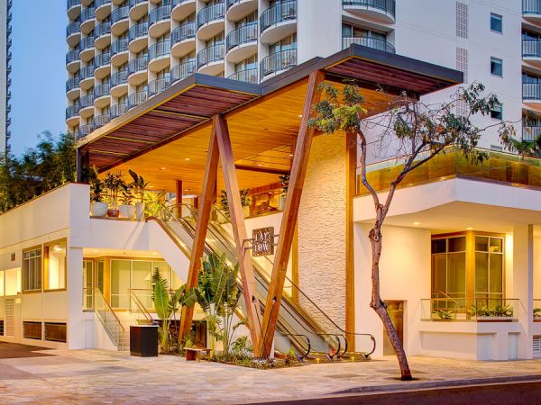 A modern building with a distinctive architectural entrance featuring wooden beams and glass elements, surrounded by greenery and urban scenery.