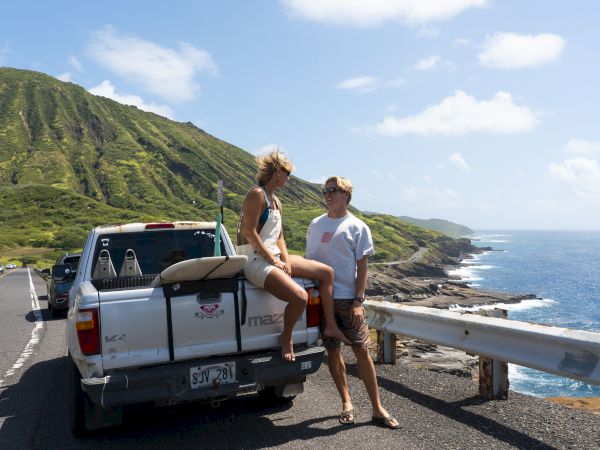 Two travelers sit on a pickup truck by the ocean highway, smiling with lush green hills and blue sky behind them.