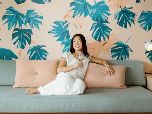 A woman in a white dress sits on a teal sofa, posing casually with beige pillows, a tropical blue-green leaf wallpaper backdrop, and a floor lamp nearby.