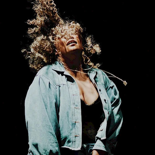 A person with curly hair wears a denim jacket and moves energetically, their hair flying against a black background.