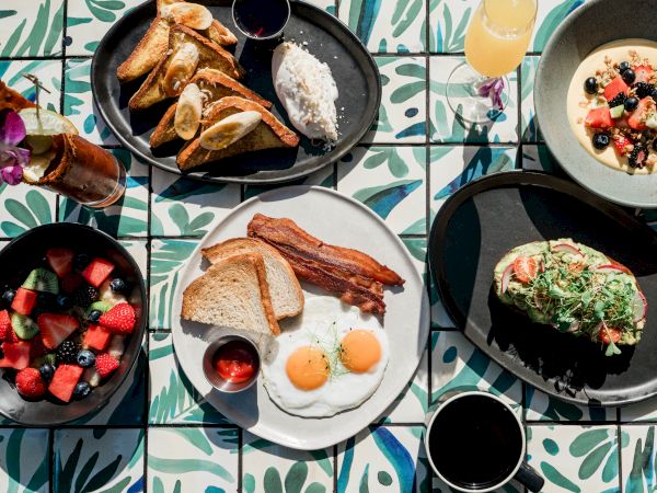 A breakfast spread with toast, eggs, bacon, fruit bowls, pancakes, and plates of avocado and granola on a colorful table, all.