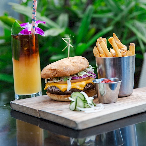 A cheeseburger with pickles, fries in a metal cup, a small sauce dish, and a tropical drink with an orchid garnish on a wooden board.