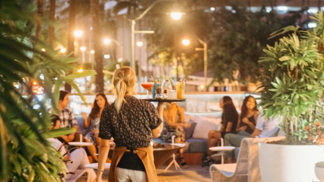 Outdoor dining scene with plants, a woman walking, and people seated at tables. Bright city lights and tall buildings in the background.