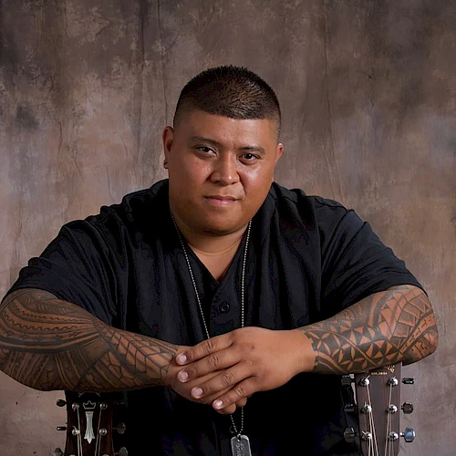 A person with tattoos sits against a textured backdrop, folding arms calmly in a studio setting.