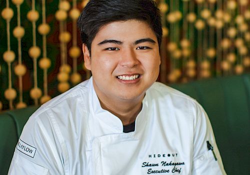 A smiling person wearing a chef's uniform with tattooed arm, sitting in a restaurant with a decorative background.