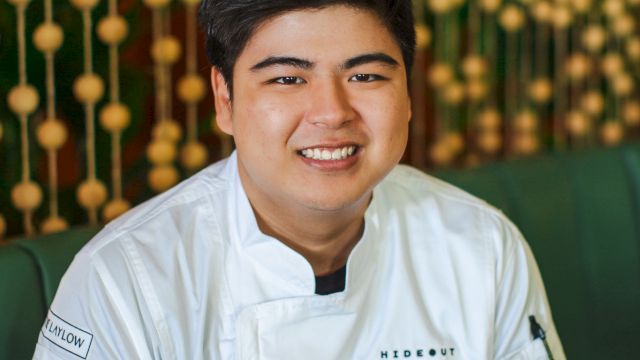 A smiling person wearing a chef's uniform with tattooed arm, sitting in a restaurant with a decorative background.