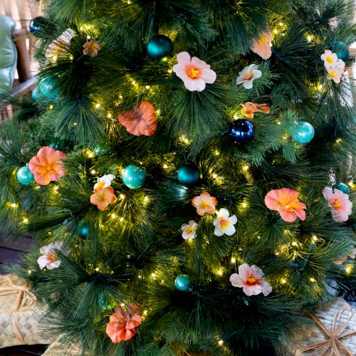 A decorated Christmas tree with green tinsel, warm lights, turquoise ornaments, and pink flowers, wrapped in woven baskets.