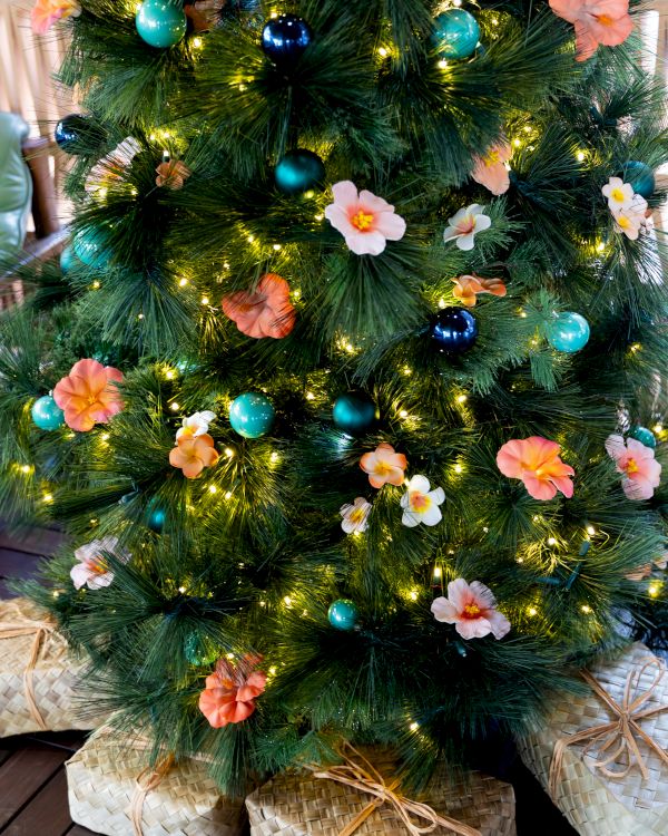 A decorated Christmas tree with green tinsel, warm lights, turquoise ornaments, and pink flowers, wrapped in woven baskets.