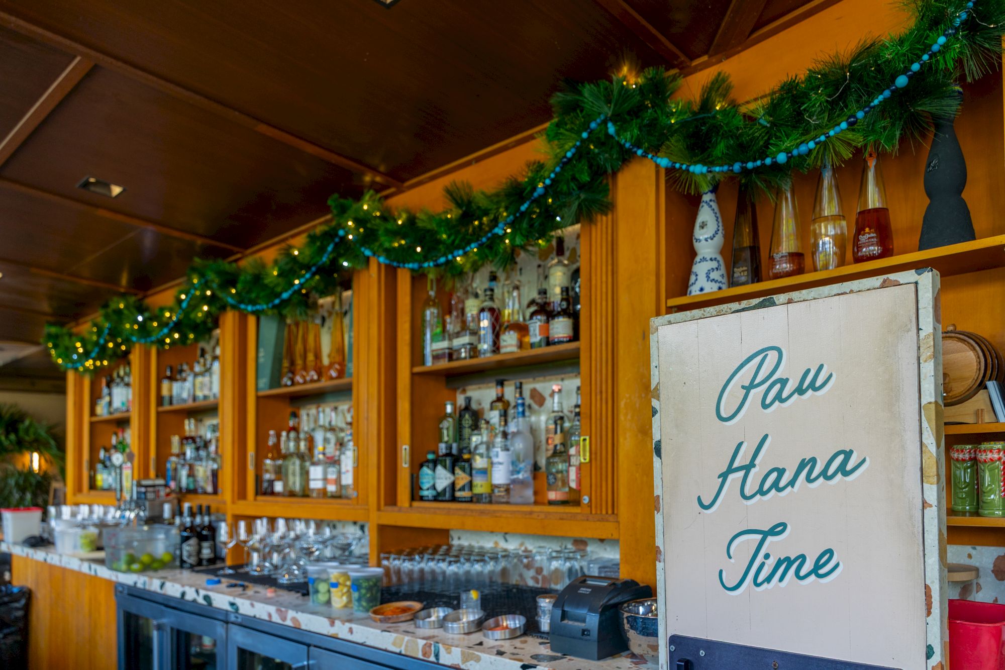 A cozy bar with bottles and holiday lights on a garland, plus a sign reading &ldquo;Paw Hana Time&rdquo;.