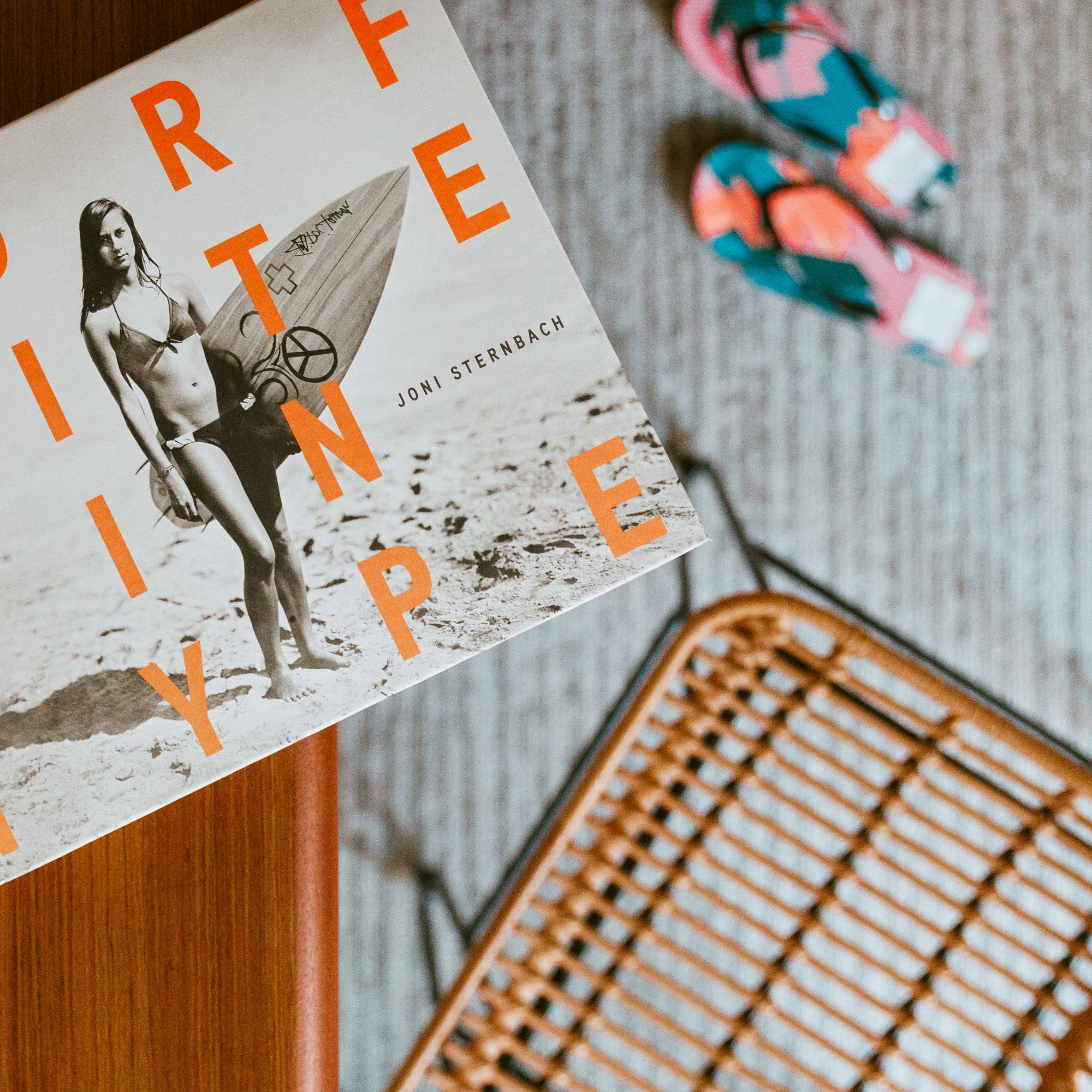 The image shows a book titled "Surf Tribe" on a table, colorful sandals on the floor, and part of a woven chair base.