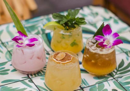 Four colorful cocktails on a decorative table, garnished with flowers, mint, and citrus slices.