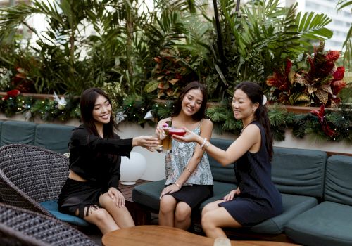 Three friends on a patio couch toast colorful drinks, smiling amid lush greenery and holiday lights.