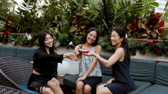 Three friends on a patio couch toast colorful drinks, smiling amid lush greenery and holiday lights.
