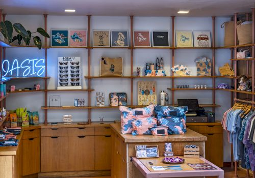 A cozy boutique with wooden shelves, colorful bags, tees, and a neon “OASIS” sign on the wall.