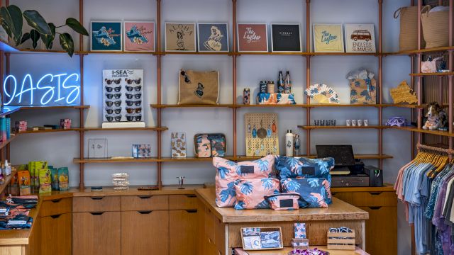 A cozy boutique with wooden shelves, colorful bags, tees, and a neon &ldquo;OASIS&rdquo; sign on the wall.