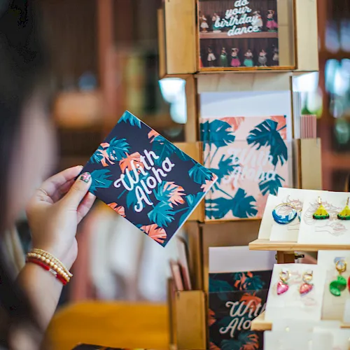 A gift shop display of printed cards and treats, with a hand holding a card reading &ldquo;With love&rdquo; and birthday posters.