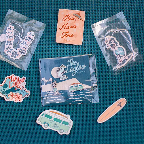 Assorted &ldquo;The Laylow&rdquo; Waikiki souvenirs&mdash;hula keychain, surfboard/van stickers, and &ldquo;Pay Hana Time&rdquo; card pieces.