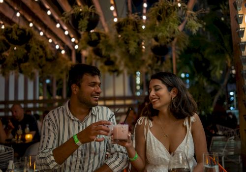Two people are at an outdoor restaurant, smiling and toasting with drinks under string lights.