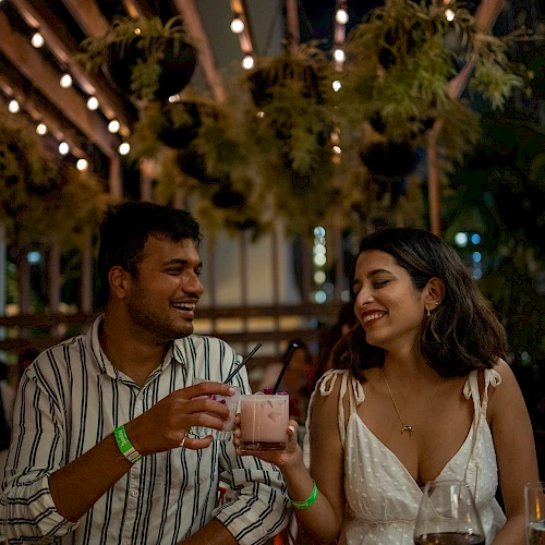 Two people are at an outdoor restaurant, smiling and toasting with drinks under string lights.