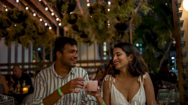 Two people are at an outdoor restaurant, smiling and toasting with drinks under string lights.