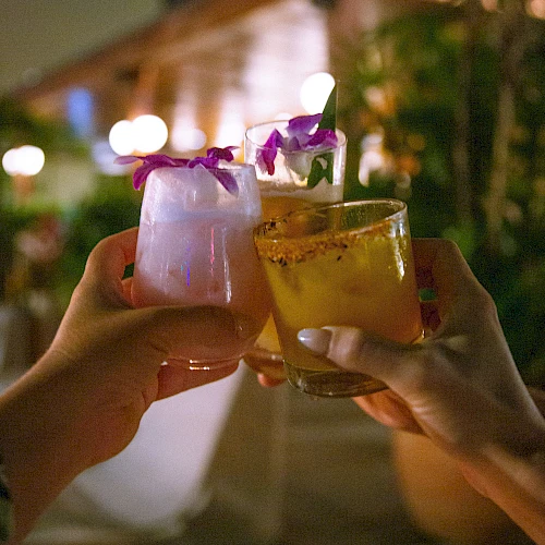 Two hands toast colorful cocktails with edible flowers in a warm, tropical-night setting. Cheers to good vibes.