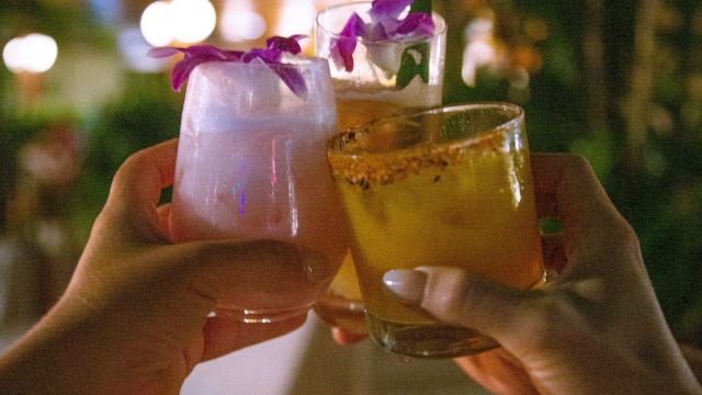 Two hands toast colorful cocktails with edible flowers in a warm, tropical-night setting. Cheers to good vibes.