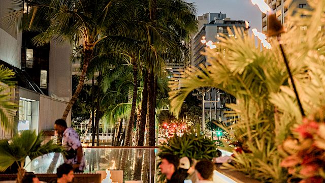 A rooftop pool scene in the city with palm trees, lounge seating, and tall buildings at dusk, lighted tiki torches adding warmth.