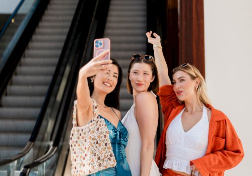 Three friends take a selfie at an escalator, posing playfully in summer outfits near a doorway with bright sunlight.