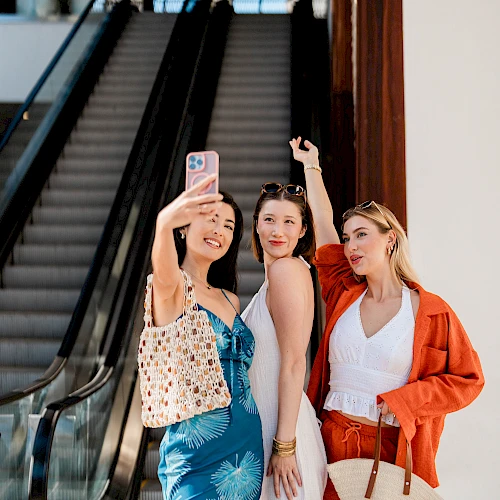 Three stylish friends pose for a selfie near an escalator, one holding up the phone, all smiling and dressed for a sunny day.