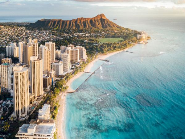 Aerial view of a coastal city with high-rise buildings along a sandy beach, crystal blue water, and Diamond Head in the distance.