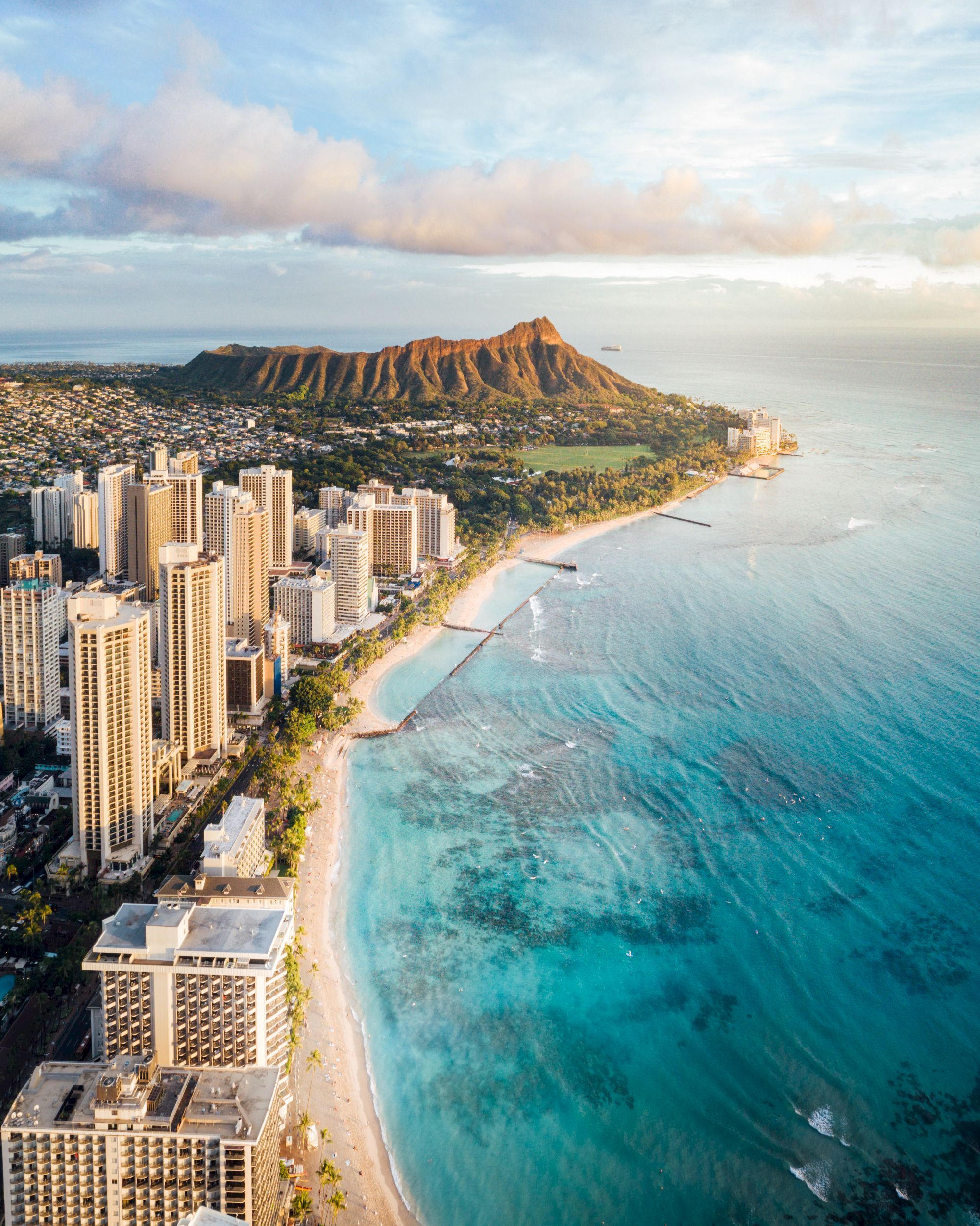 Aerial view of Honolulu: city high-rises along a sandy coastline with crystal-blue waters, Diamond Head in the distance, vibrant tropical scenery.