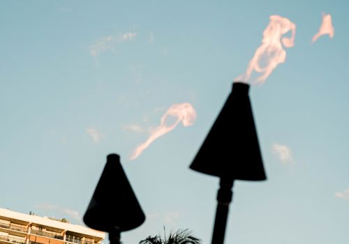 A skyline with tall modern buildings and a palm tree, foregrounded by two torches with flames against a clear blue sky.