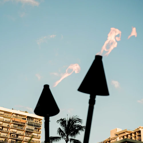 A skyline with tall modern buildings and a palm tree, foregrounded by two torches with flames against a clear blue sky.