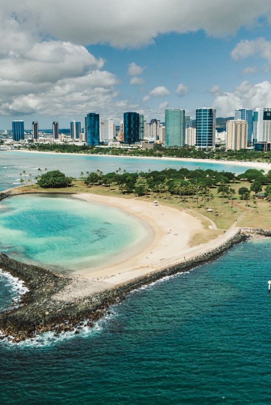 A tropical coastal scene with a small curved beach, turquoise lagoon, and a modern city skyline in the distance, framed by blue seas and clouds.