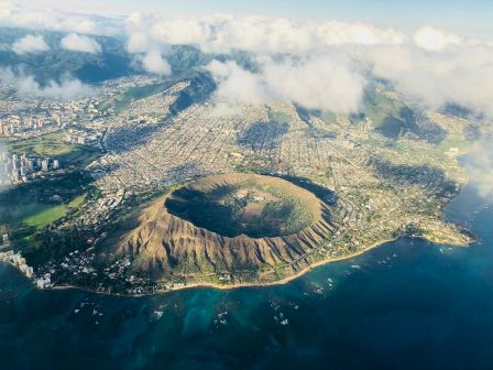 Aerial view of a volcanic crater lake city built around a circular volcanic cone, with coastlines, buildings, and clouds above.